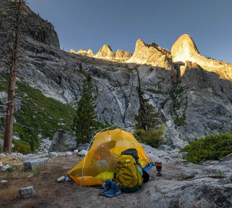 A bright yellow tent set up on rocky terrain in the mountains, with a backpack and hiking boots outside, surrounded by steep granite cliffs and sunlit peaks in the background.