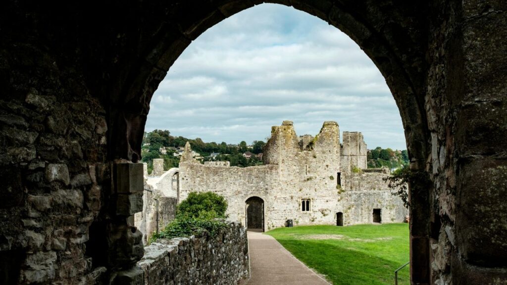 View through a stone archway of ancient castle ruins, with weathered walls, grassy grounds, and a backdrop of distant hills under a cloudy sky.