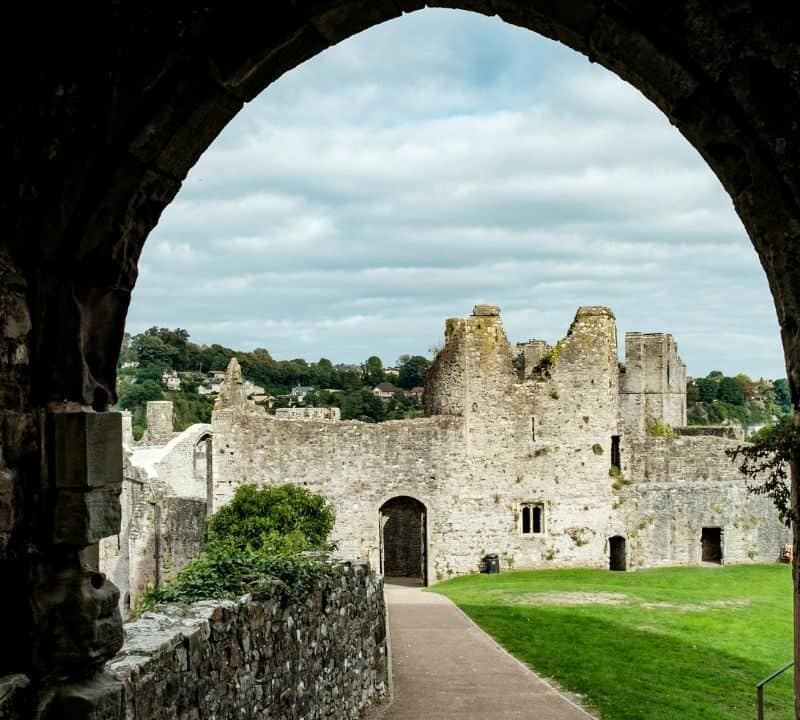 View through a stone archway of ancient castle ruins, with weathered walls, grassy grounds, and a backdrop of distant hills under a cloudy sky.