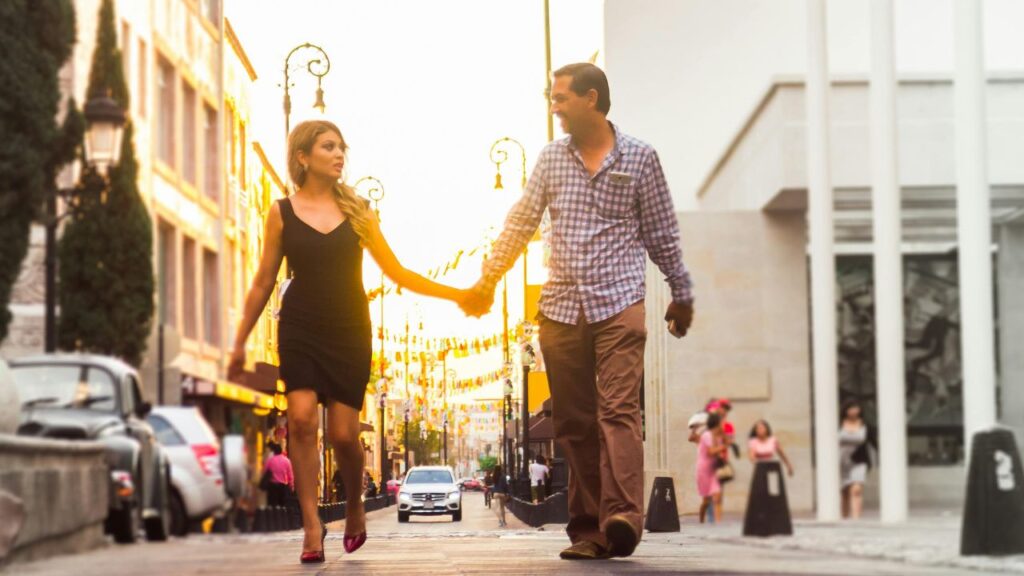 A couple holding hands and walking down a sunlit city street, surrounded by historic buildings, cars, and street lamps.