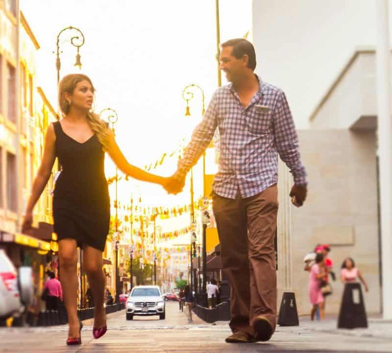 A couple holding hands and walking down a sunlit city street, surrounded by historic buildings, cars, and street lamps.