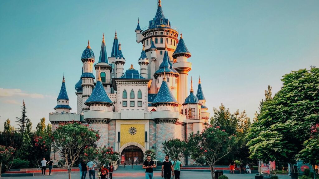 A fairy-tale style castle with blue turrets and towers surrounded by trees, with visitors walking in the foreground under a clear sky.