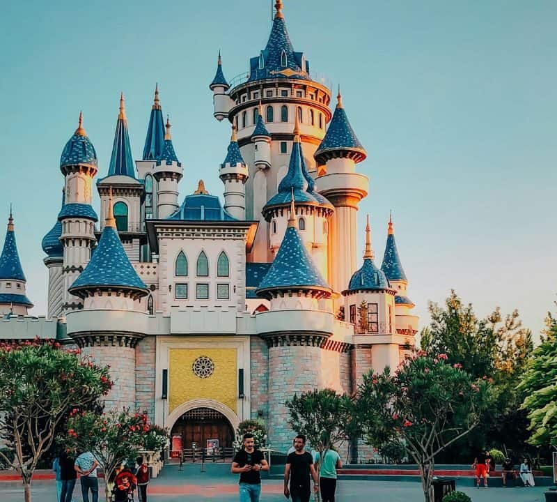 A fairy-tale style castle with blue turrets and towers surrounded by trees, with visitors walking in the foreground under a clear sky.