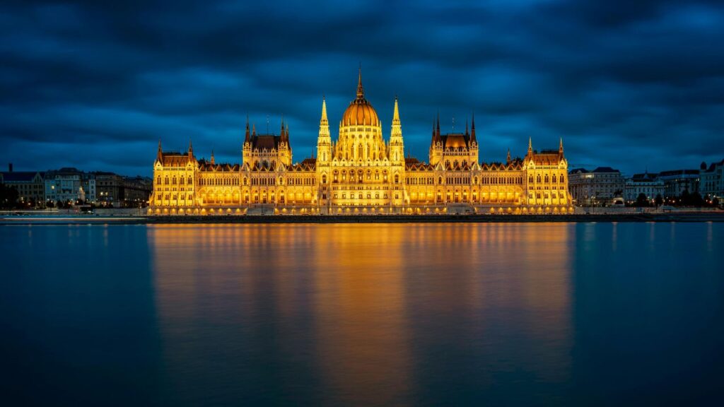 The illuminated Hungarian Parliament Building at night, reflecting on the calm waters of the Danube River in Budapest under a dramatic cloudy sky.