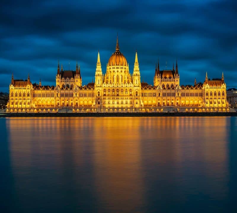 The illuminated Hungarian Parliament Building at night, reflecting on the calm waters of the Danube River in Budapest under a dramatic cloudy sky.