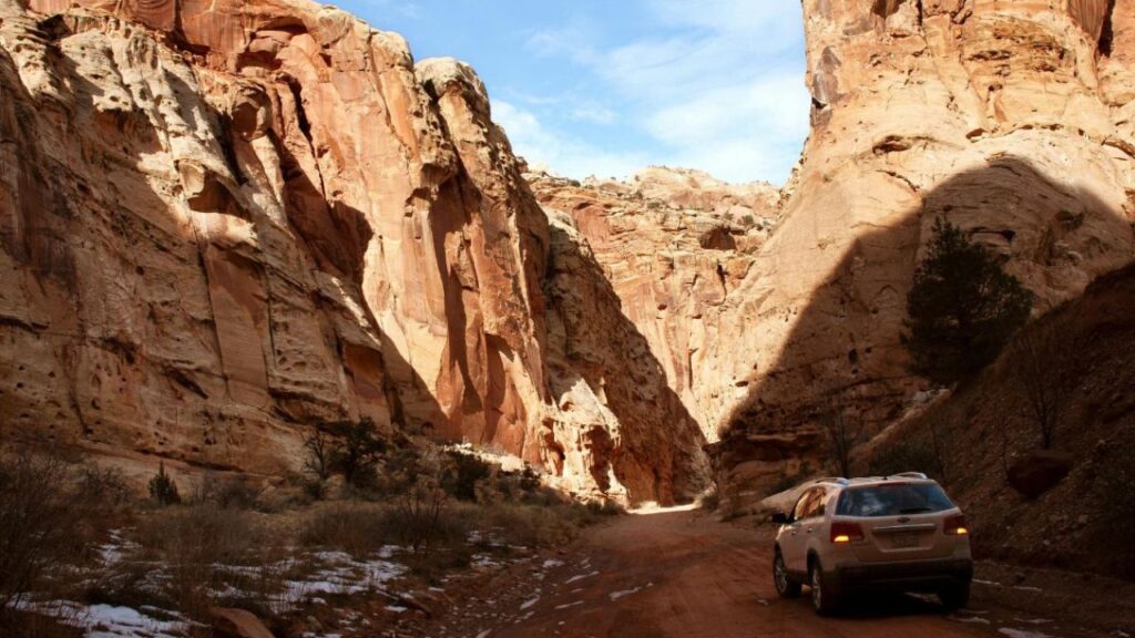 A white SUV drives through a narrow canyon with towering red rock cliffs and patches of snow on the ground under a partly cloudy sky.