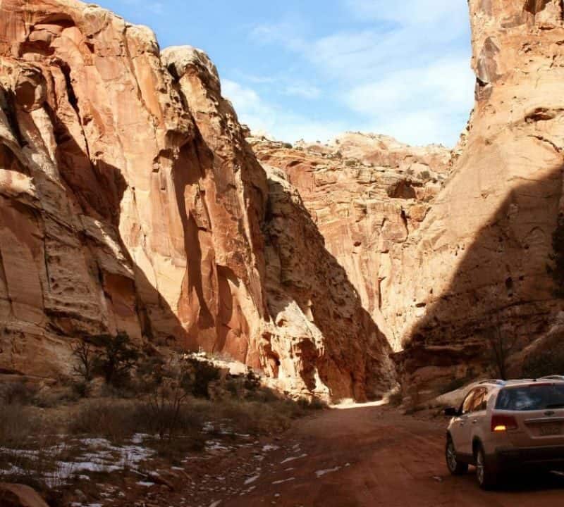 A white SUV drives through a narrow canyon with towering red rock cliffs and patches of snow on the ground under a partly cloudy sky.