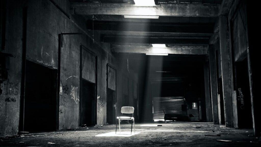 A single chair sits under a beam of light in an abandoned, graffiti-covered building, creating a haunting and eerie atmosphere.