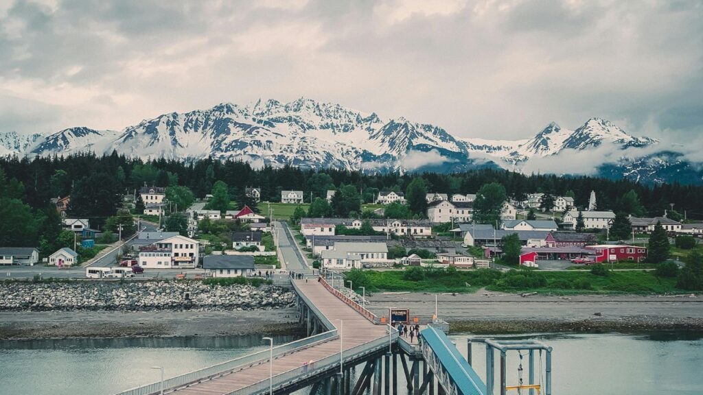 Scenic view of Haines, Alaska, with a long wooden pier leading into town, colorful houses surrounded by trees, and snow-capped mountains in the background under a cloudy sky.