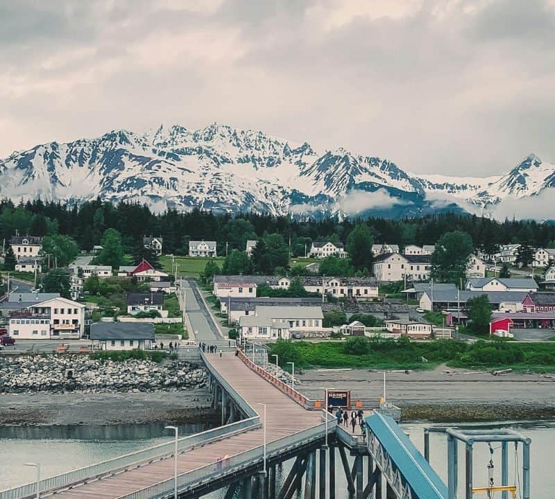 Scenic view of Haines, Alaska, with a long wooden pier leading into town, colorful houses surrounded by trees, and snow-capped mountains in the background under a cloudy sky.