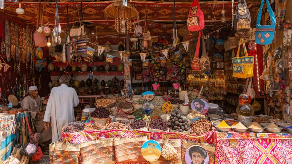 : A vibrant traditional market stall filled with colorful spices, dried fruits, woven bags, handcrafted goods, and hanging decorations, with two men conversing inside.
