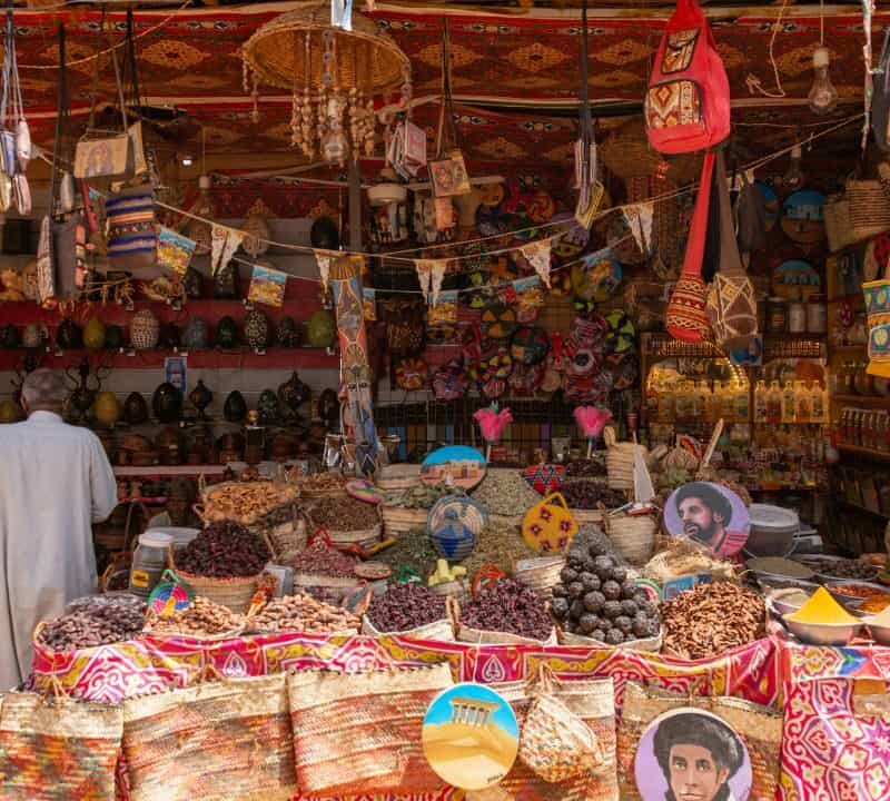 : A vibrant traditional market stall filled with colorful spices, dried fruits, woven bags, handcrafted goods, and hanging decorations, with two men conversing inside.