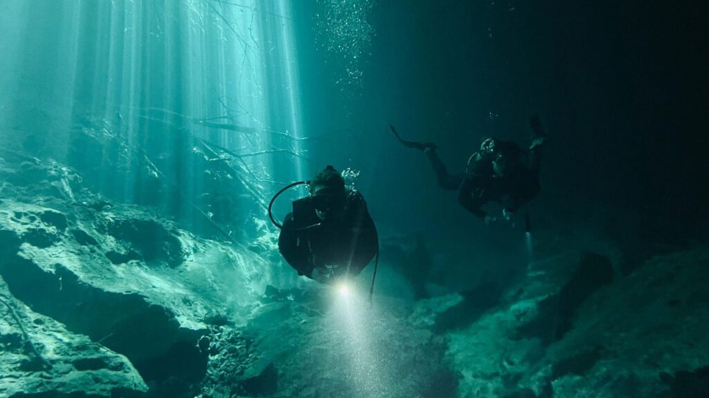 Two scuba divers explore a dark underwater cave, illuminated by beams of sunlight filtering through the water and flashlights highlighting rocky terrain.