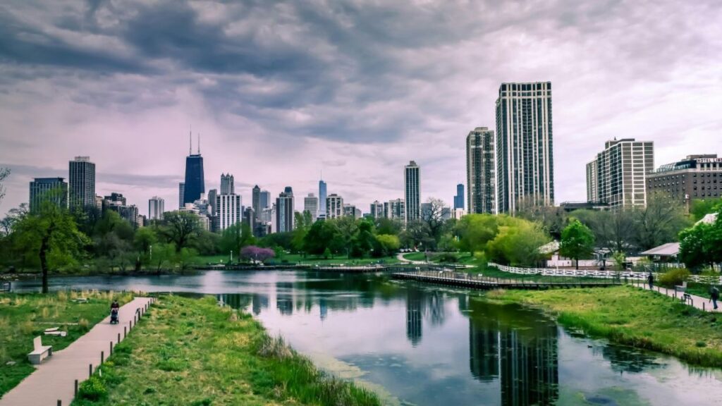 Skyline view of Chicago with tall buildings reflected in a calm pond at Lincoln Park, surrounded by green trees and walking paths.