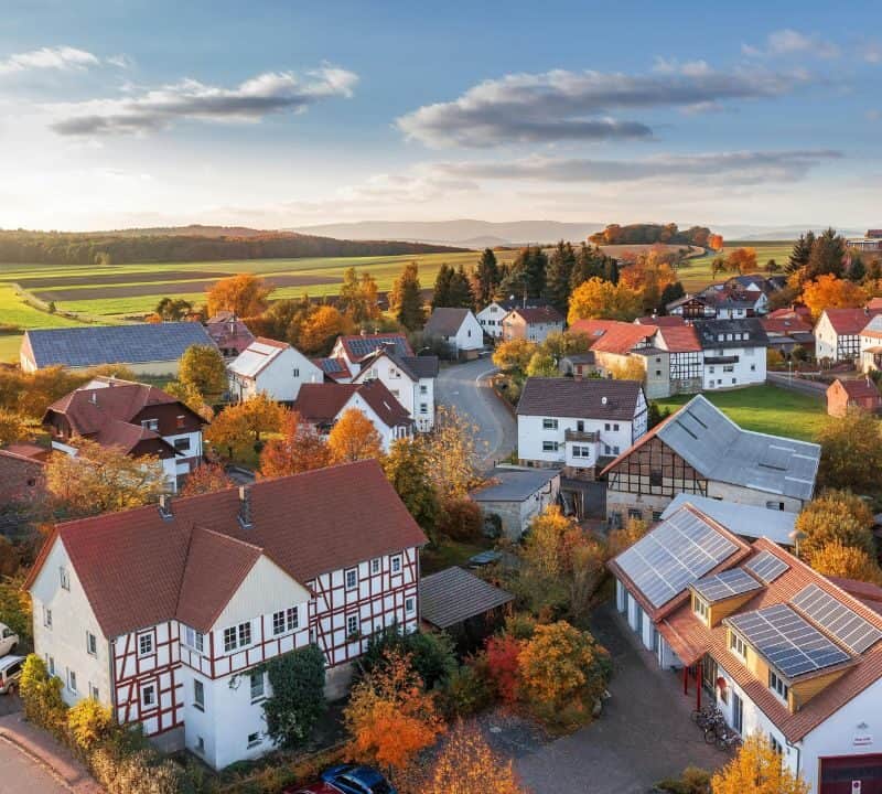Aerial view of a charming small town surrounded by fields and rolling hills, with red-roofed houses, autumn foliage, and a peaceful rural landscape at sunset.