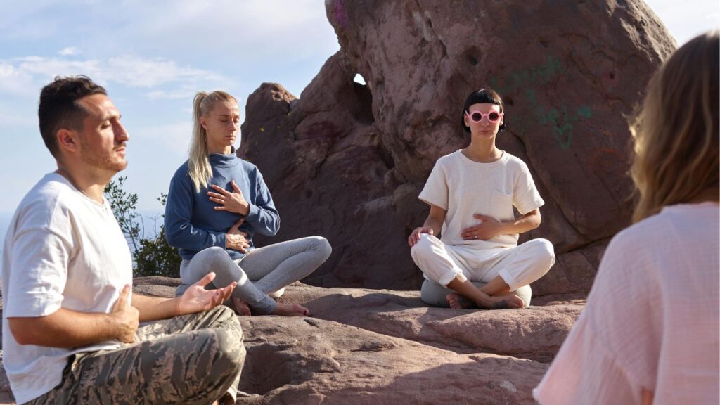 A small group of people meditating outdoors on rocky terrain, sitting cross-legged with eyes closed and hands on their chests or laps, under natural sunlight.