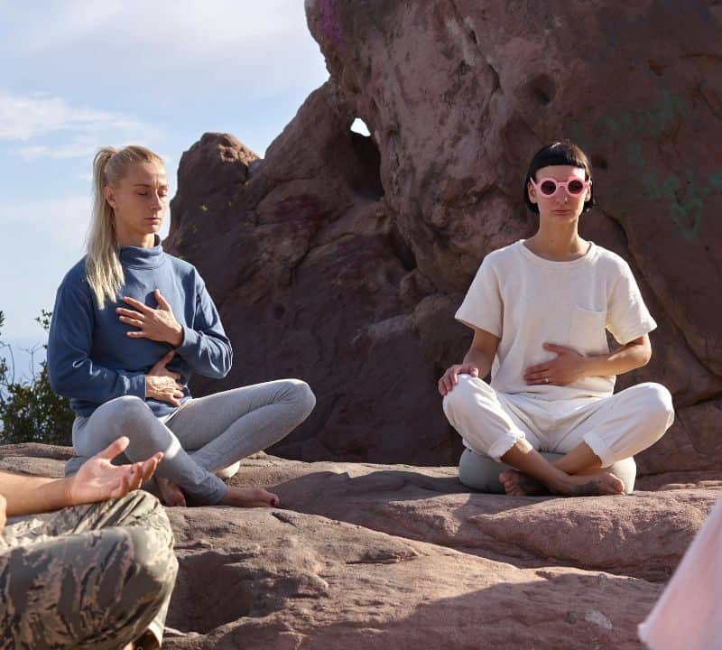 A small group of people meditating outdoors on rocky terrain, sitting cross-legged with eyes closed and hands on their chests or laps, under natural sunlight.