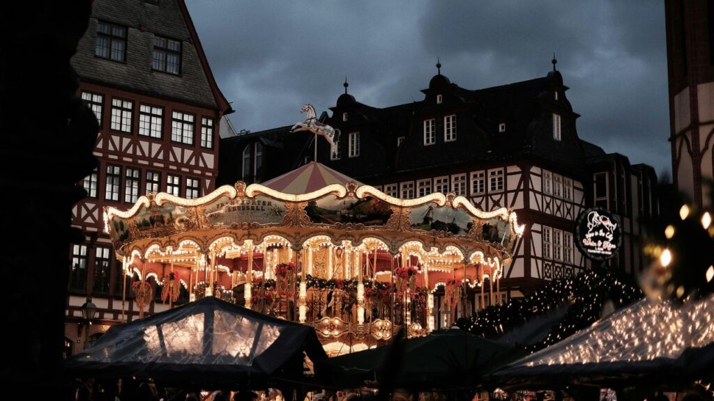 Illuminated vintage carousel at night in a town square, surrounded by traditional half-timbered buildings and market stalls decorated with lights.