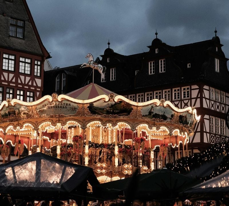 Illuminated vintage carousel at night in a town square, surrounded by traditional half-timbered buildings and market stalls decorated with lights.