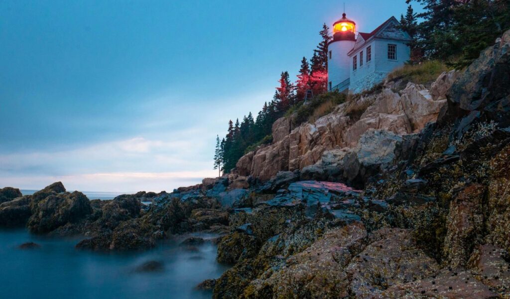 A glowing lighthouse stands on a rocky cliff surrounded by pine trees at dusk in Acadia National Park, Maine, overlooking the Atlantic Ocean.