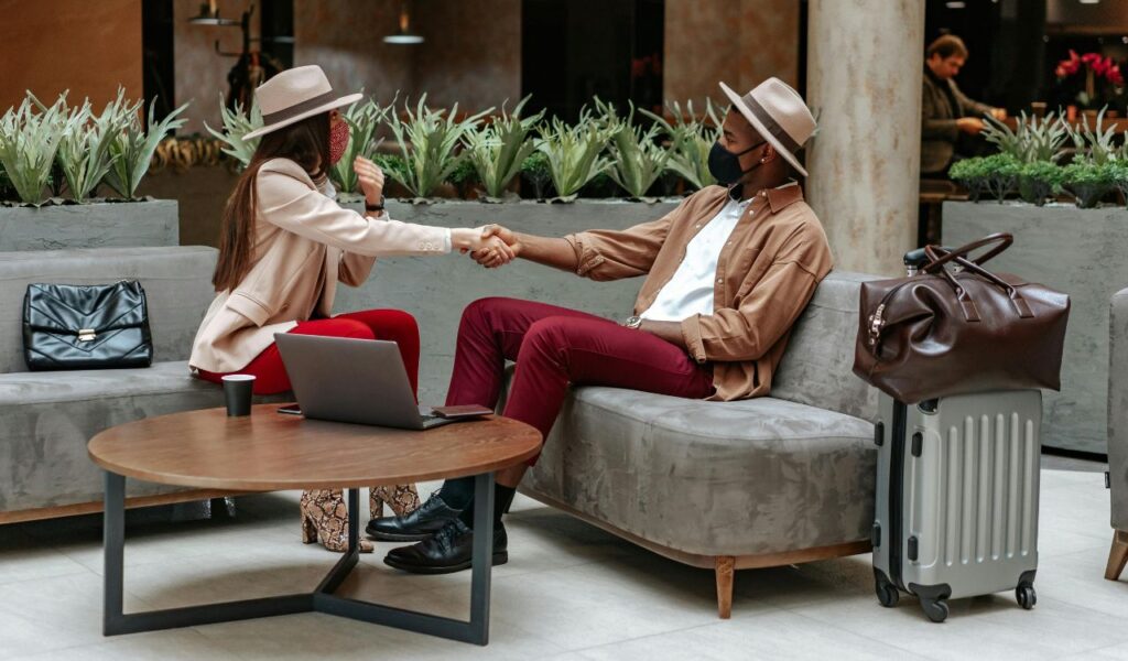 Two travelers wearing face masks and hats shake hands while seated in an airport lounge, with luggage, a laptop, and bags nearby.