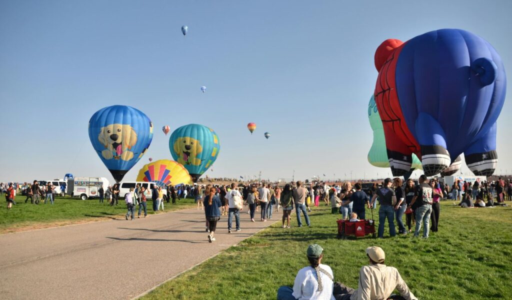 Crowds at a hot air balloon festival in Albuquerque, New Mexico, with colorful balloons shaped like dogs and superheroes rising into a clear blue sky.