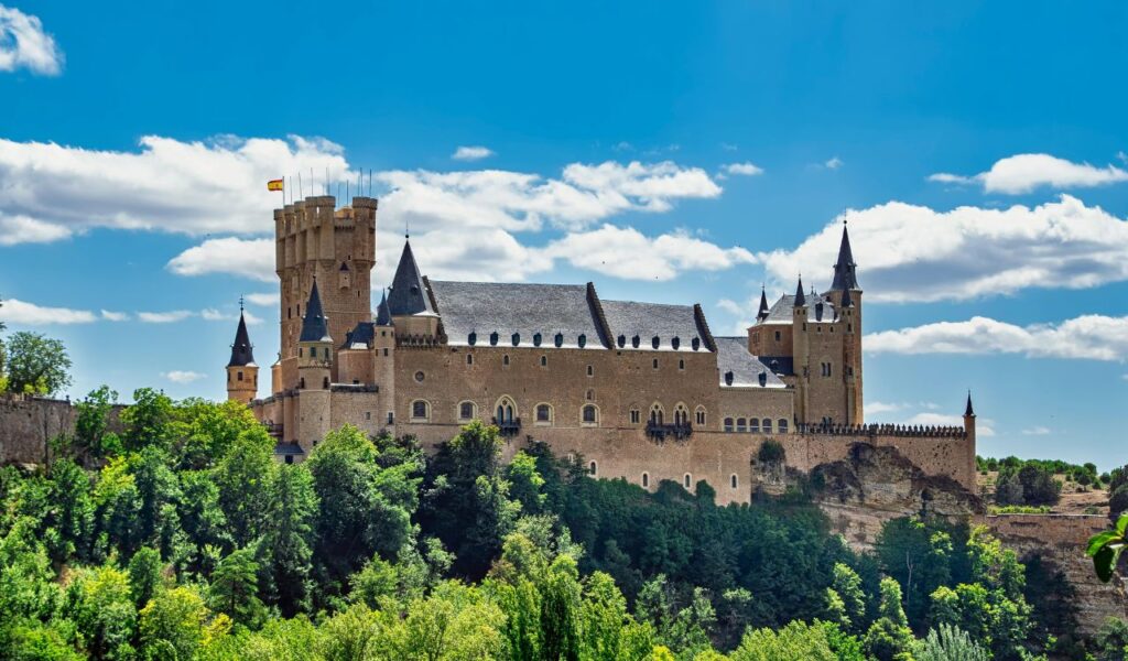 Alcázar of Segovia in Spain, a medieval castle with tall towers and a Spanish flag, perched on a rocky hill surrounded by lush green trees under a bright blue sky.