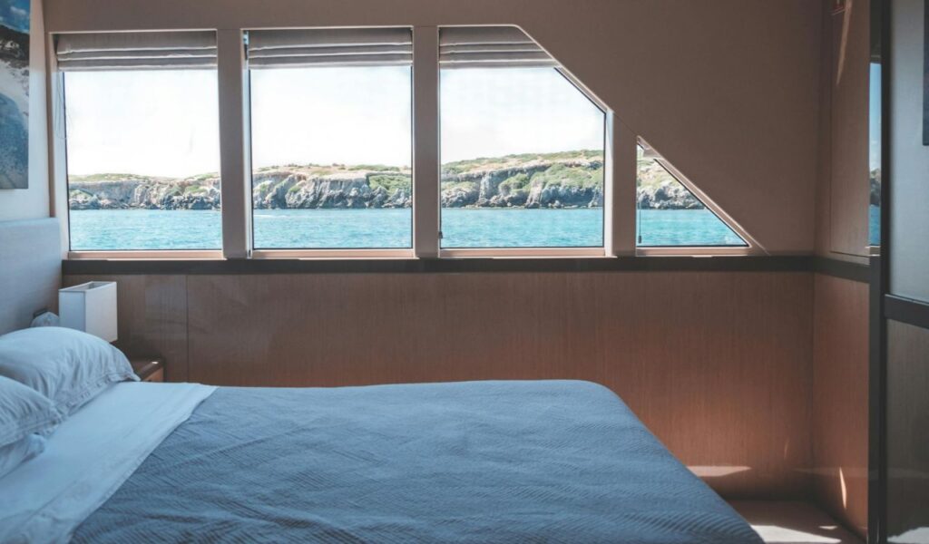 A cozy cruise ship cabin with a neatly made bed and large windows offering a scenic ocean view with rocky coastline in the distance.