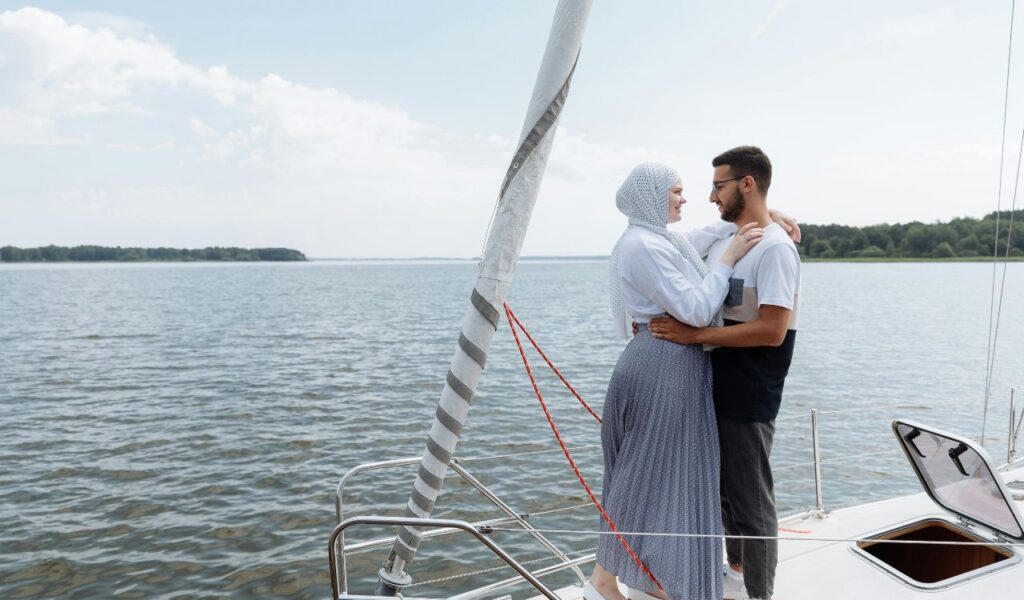 A couple stands close together on the deck of a sailboat, embracing while looking into each other’s eyes, with calm water and distant trees in the background.