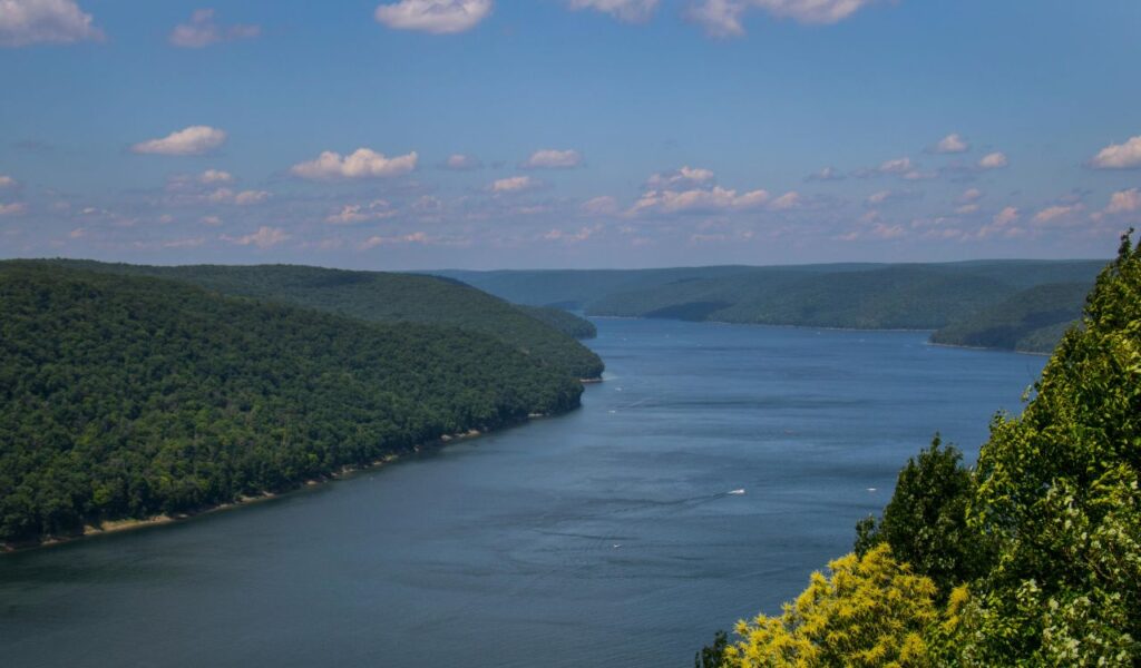 A wide, calm river winds through lush green forested hills under a partly cloudy sky in Allegheny, New York