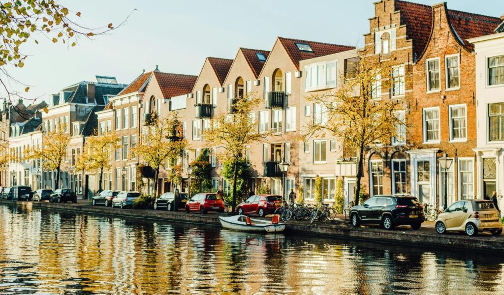 A peaceful Amsterdam canal lined with traditional Dutch houses, bicycles, and parked cars, with autumn trees reflecting in the golden water.