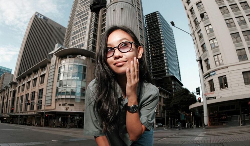 A young woman wearing glasses and a watch poses thoughtfully in an urban setting surrounded by tall modern and historic buildings.