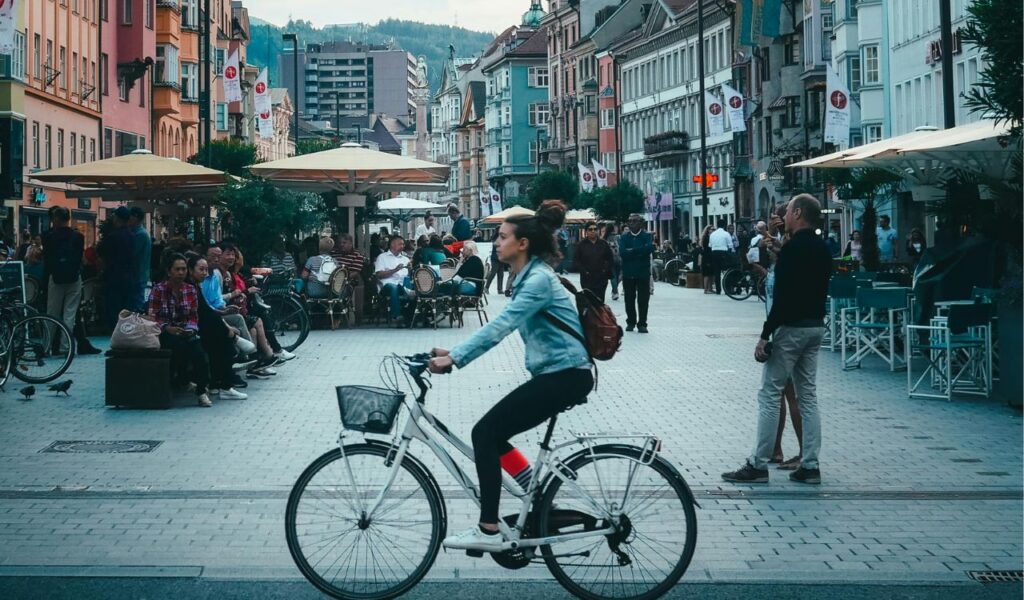 ChatGPT said:
A woman in Austria rides her bicycle through a lively pedestrian street lined with colorful buildings, outdoor cafés, and people enjoying the day.