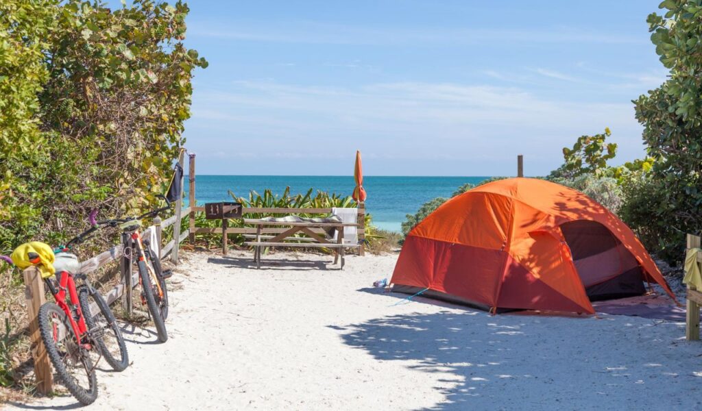 A bright orange tent sits on a sandy campsite by the ocean, with two bicycles leaning against a wooden fence and a picnic table nearby.