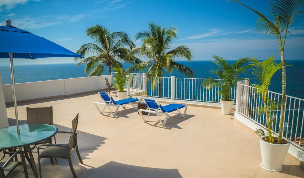 Spacious cruise ship balcony with blue lounge chairs, palm trees in white pots, and a glass table under a blue umbrella overlooking the ocean.
