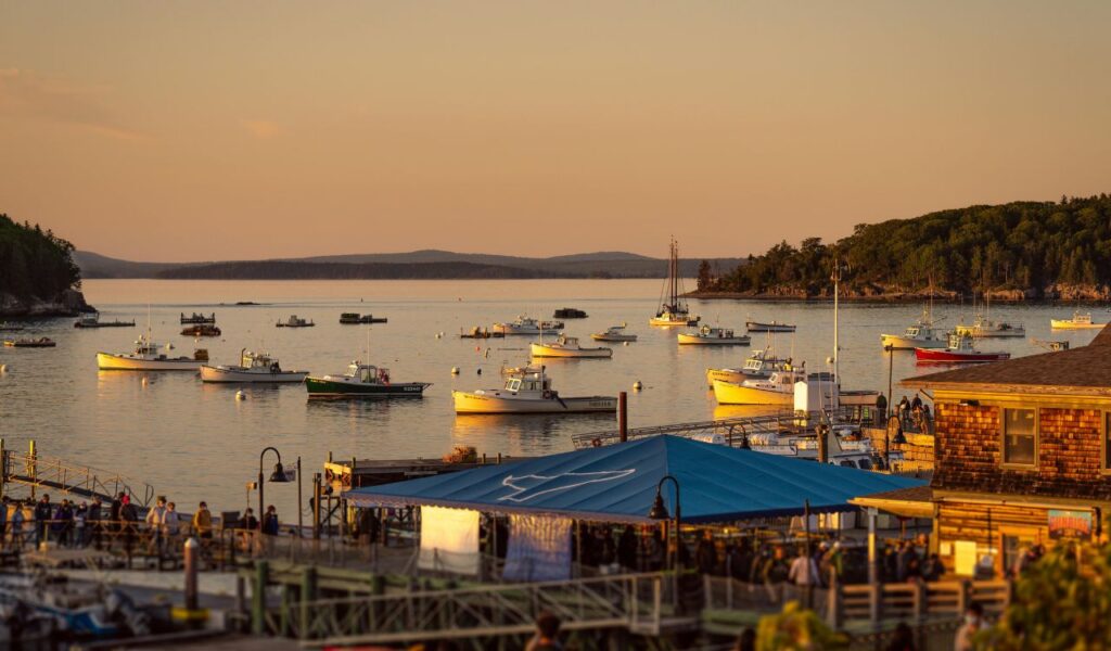 Boats anchored in the calm waters of Bar Harbor, Maine at sunset, with golden light reflecting on the water, forested hills in the background, and a busy waterfront area in the foreground.