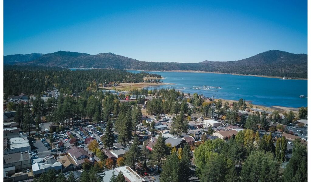 Aerial view of Big Bear Lake surrounded by forested hills, residential buildings, and a marina with boats under a clear blue sky.