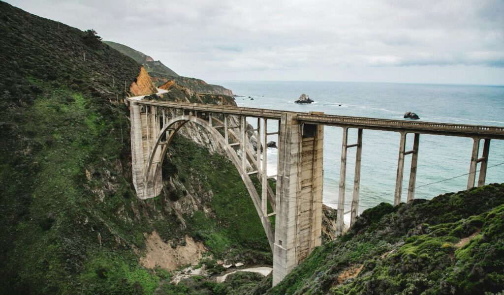 Bixby Creek Bridge in Big Sur, California, stretching over a rugged canyon with lush green hills and the Pacific Ocean in the background.