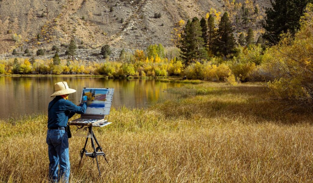 An artist painting a scenic autumn landscape by a calm lake in Bishop, California, surrounded by golden grass, colorful trees, and distant rocky hills