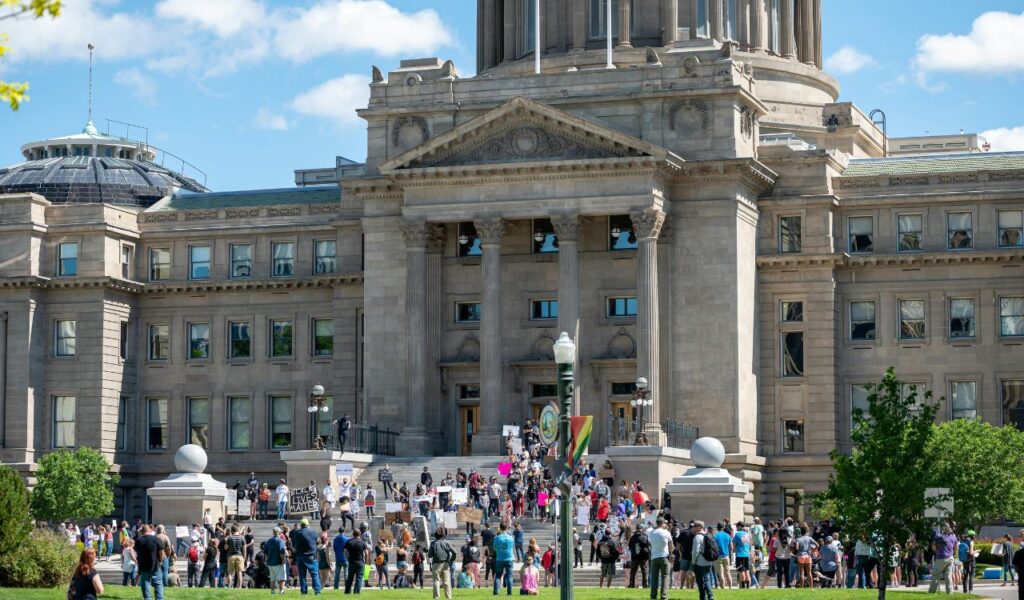 Crowd gathered in front of the Idaho State Capitol in Boise for a peaceful protest on a sunny day, with people holding signs on the building steps