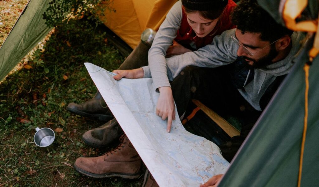A couple sitting inside a tent examines a large paper map together, with one person pointing to a location, suggesting travel planning or navigation during a camping trip.