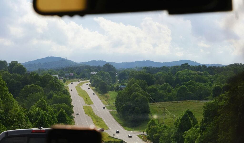 Scenic highway view in Boone, North Carolina, surrounded by lush green trees and rolling hills under a partly cloudy sky, with cars driving along the road and distant mountains in the background
