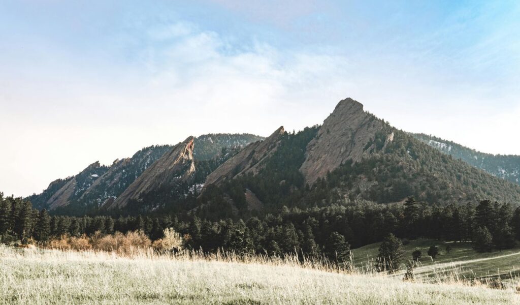 Panoramic view of the Flatirons rock formations in Boulder, Colorado, with forested slopes and grassy meadows under a soft blue sky.