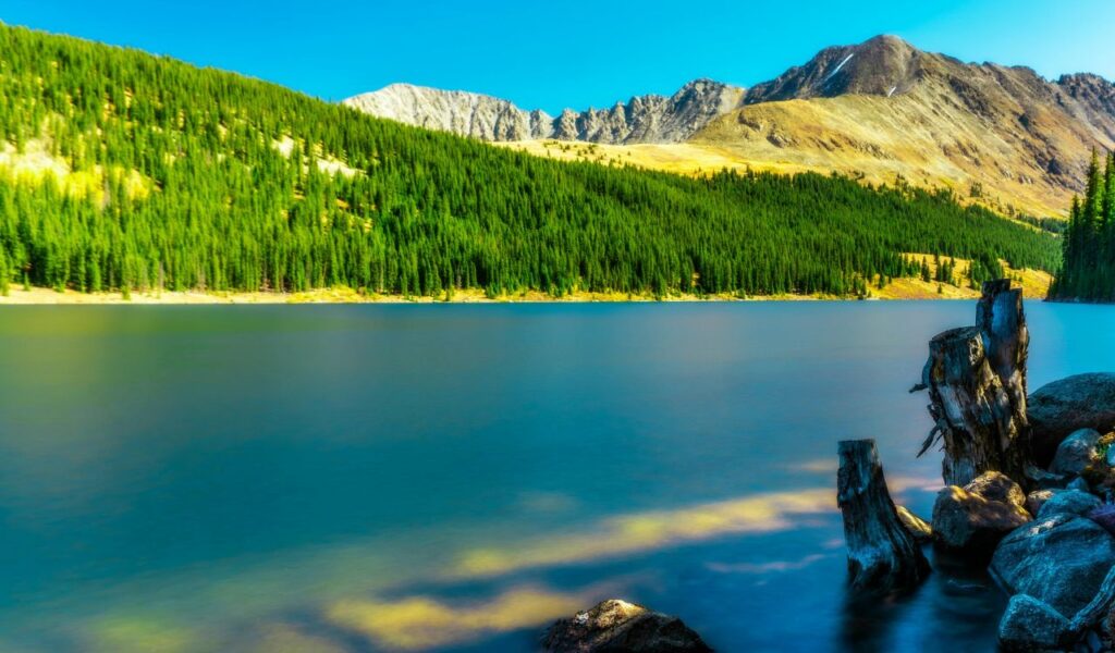 Crystal-clear alpine lake near Breckenridge, Colorado, surrounded by dense pine forests and rugged mountain peaks under a bright blue sky.