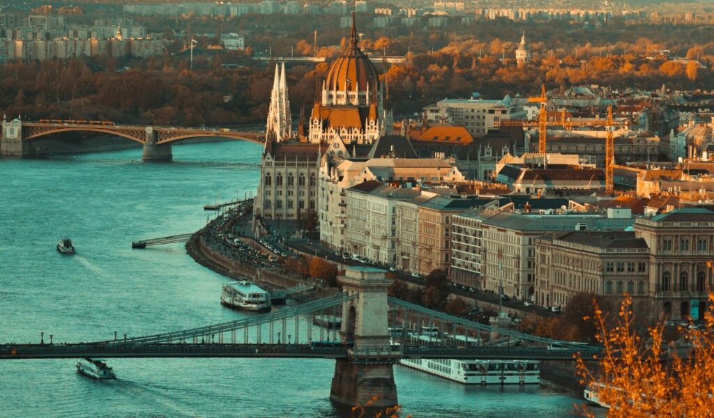 View of Budapest’s Parliament Building along the Danube River, with bridges, boats, and the city bathed in warm golden light.