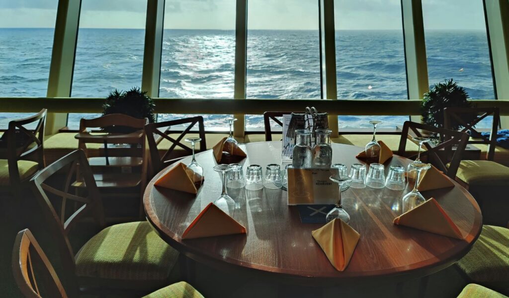 Dining table set with glasses and napkins by large cruise ship windows overlooking the sea.