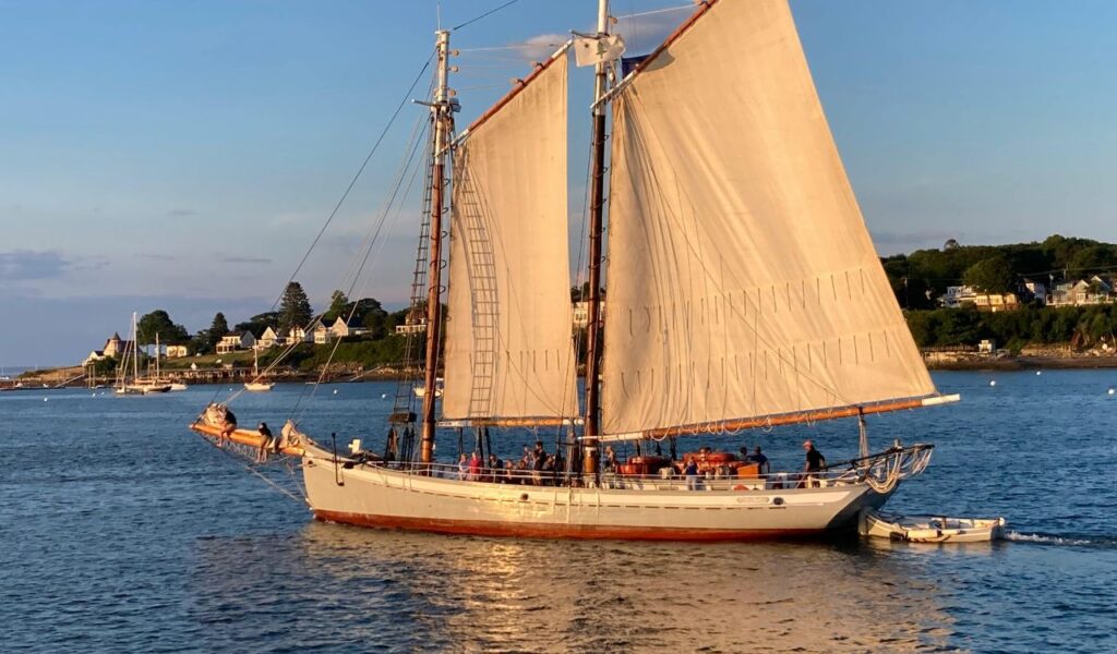 A classic two-masted sailboat with white sails glides near the coast of Camden Hills, Maine, during golden hour, with shoreline houses and trees in the background.