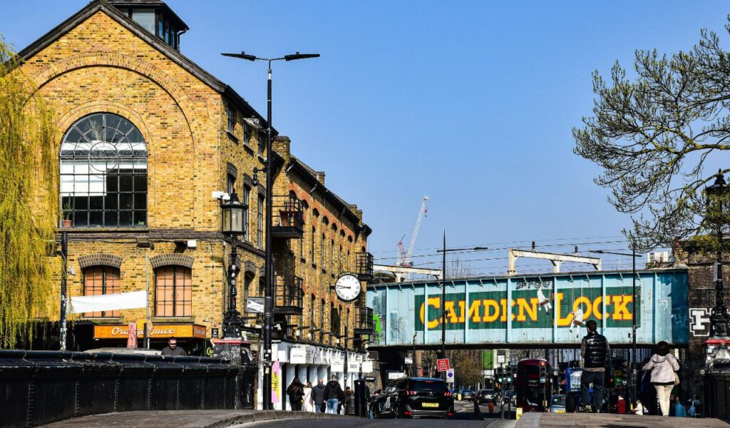 Camden, London street with the iconic blue Camden Lock bridge, brick buildings, shops, and pedestrians on a sunny day.