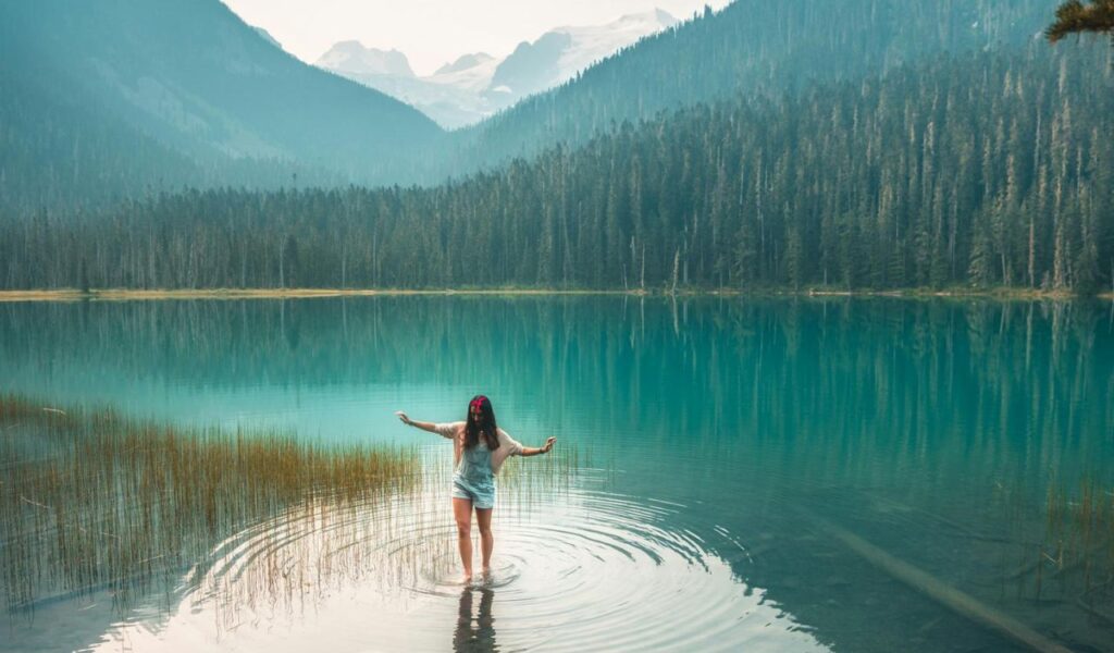 A woman stands in the shallow waters of a crystal-clear lake in Canada, surrounded by pine forests and distant snow-capped mountains.