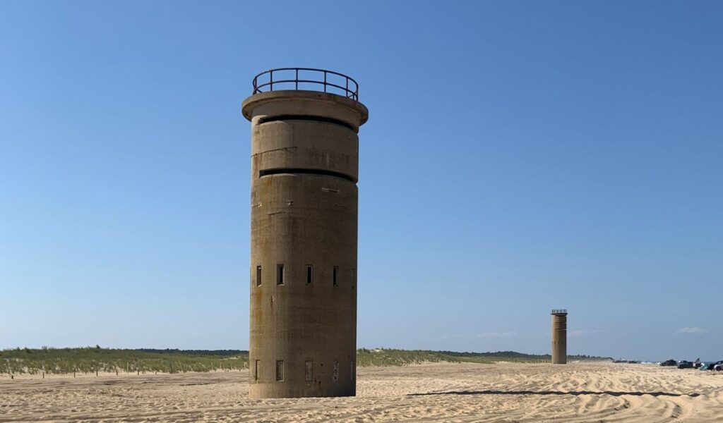 Two historic World War II observation towers stand on the sandy shoreline of Cape Henlopen State Park under a clear blue sky.